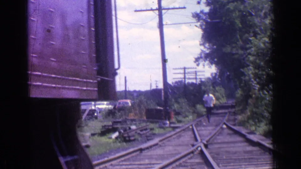 1976 Chicago trackside scene with switch rails, utility poles, parked cars, trees, and a distant person walking near the tracks.