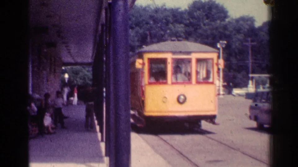 Yellow trolley approaching a Chicago station platform in 1976, with seated passengers, tracks in pavement, trees, and a parked car nearby.