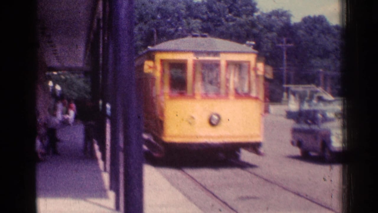 Yellow trolley car approaches a covered platform with pedestrians and cars nearby.
