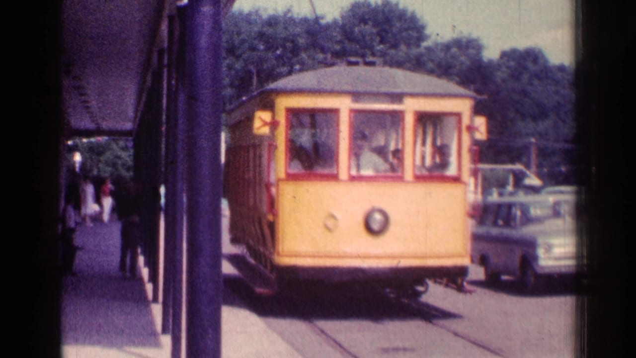 1976 Chicago 8mm frame of a yellow trolley-style car nearing the stop, with passengers visible inside and a parked car beside it.