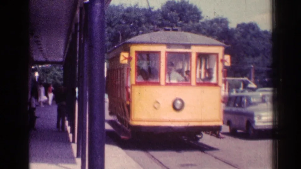 1976 Chicago 8mm frame of a yellow trolley-style car nearing the stop, with passengers visible inside and a parked car beside it.