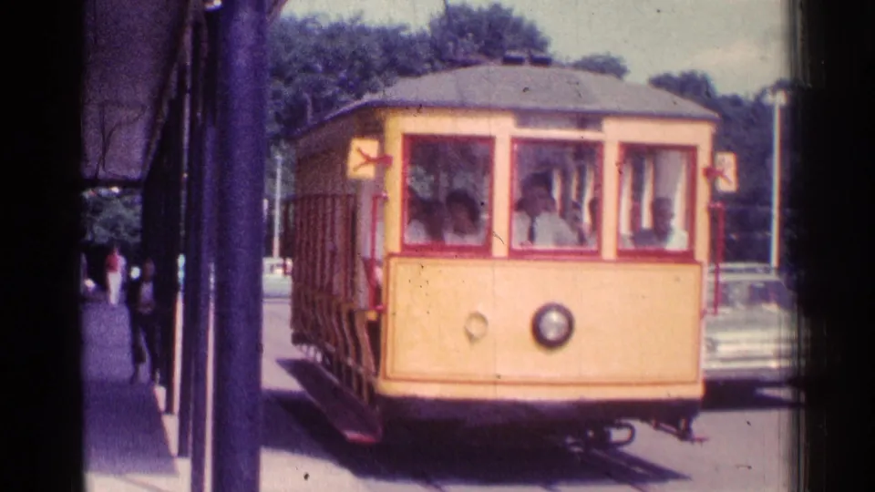 1976 Chicago 8mm frame showing the trolley nearly at the platform, with seated passengers behind large front windows and a car at the curb.