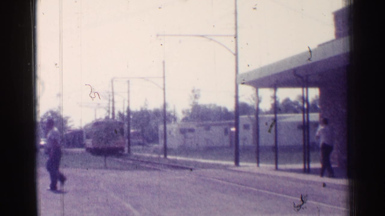 Trolley nears the stop while pedestrians stand near the track and canopy.