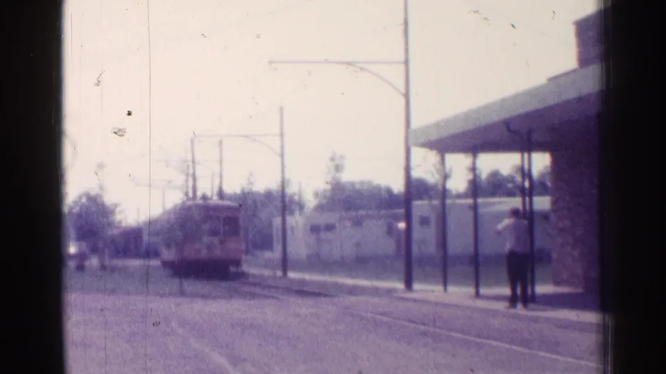 Chicago 1976 home-movie frame with the trolley on street tracks, overhead wires, and a man waiting near the canopy at right.