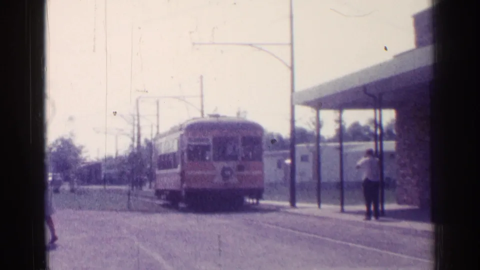 1976 Chicago 8mm frame showing a trolley-like transit car approaching a street stop with overhead wires and a waiting adult.