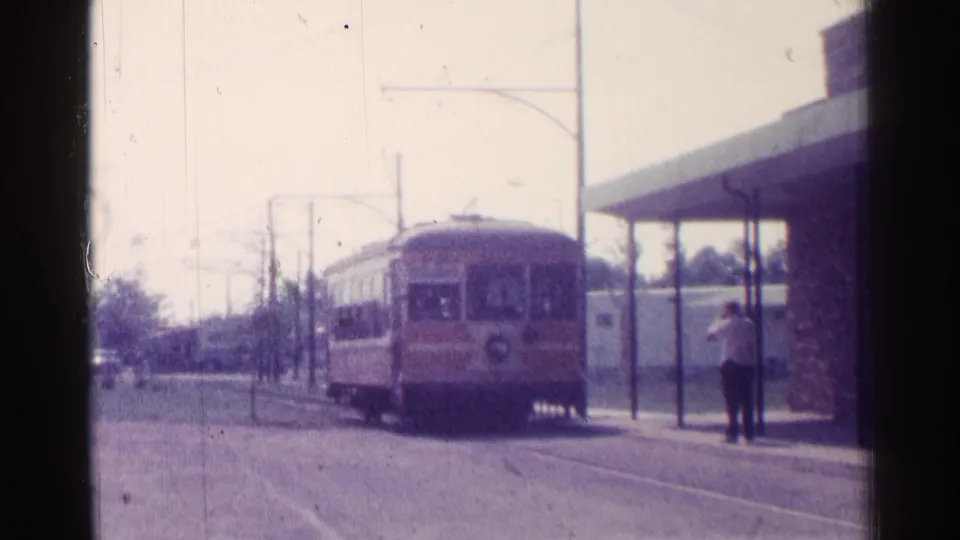 The trolley-like vehicle moves closer to the stop beside a simple shelter, with overhead wires and street poles visible.