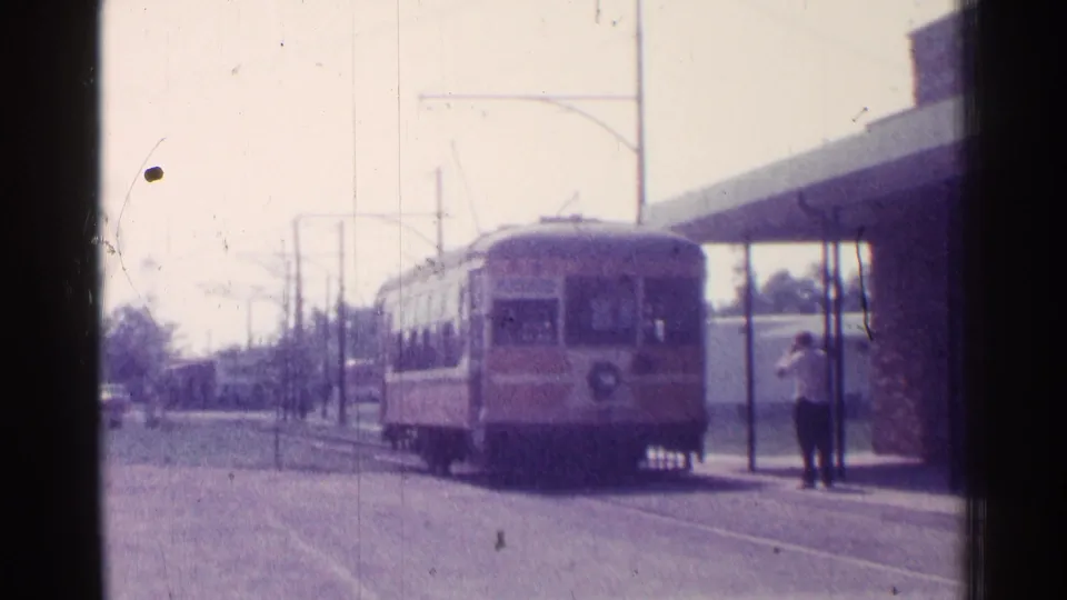 A soft 8mm frame shows the transit car advancing along the street toward a waiting adult at the shelter.