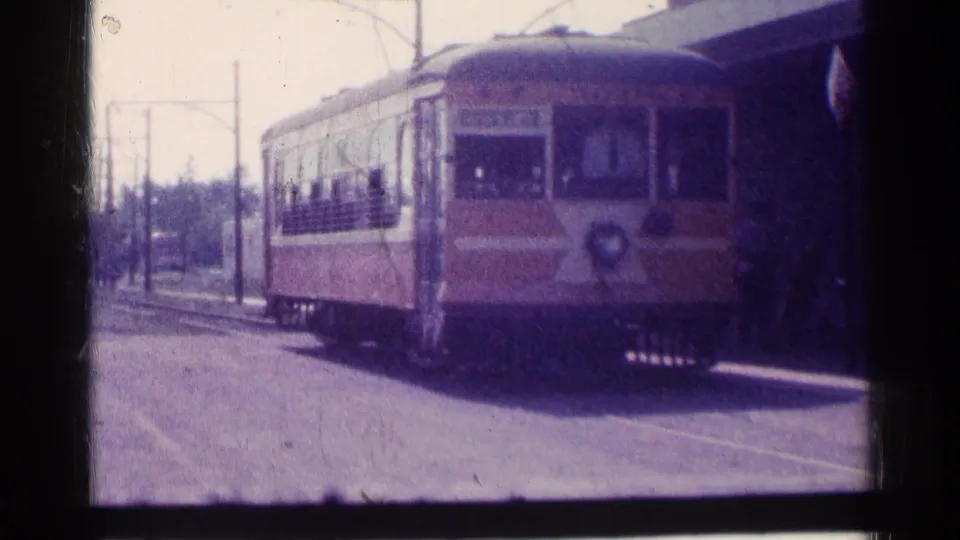 The trolley-like car fills more of the frame as it reaches the stop area, with windows, overhead wires, and shelter posts visible.