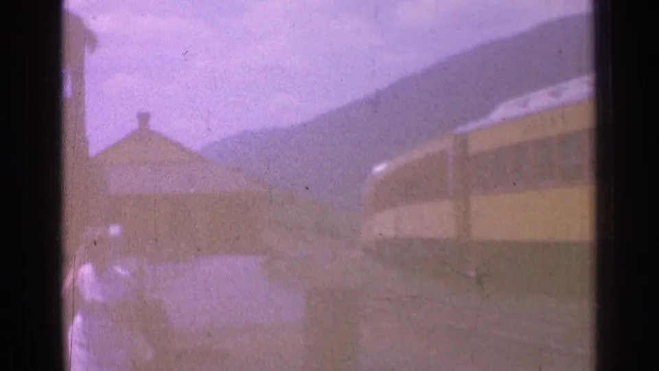 Two distant people pass through a hazy 1976 Chicago rail yard near a long yellow train and low industrial buildings.