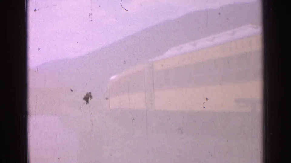 A long yellow train and low buildings sit in a hazy Chicago rail-yard view, softened by aged 8mm film grain and scratches.