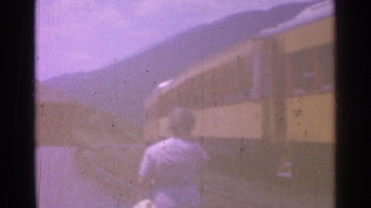 A woman in a light dress walks beside yellow train cars in the 1976 home-movie footage, with hills in the background.