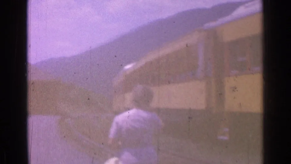 A woman in a light dress walks beside yellow train cars in the 1976 home-movie footage, with hills in the background.