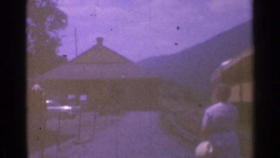 Handheld 8mm frame shows a woman near yellow train cars, a small station building, parked cars, hills, and bright sky.
