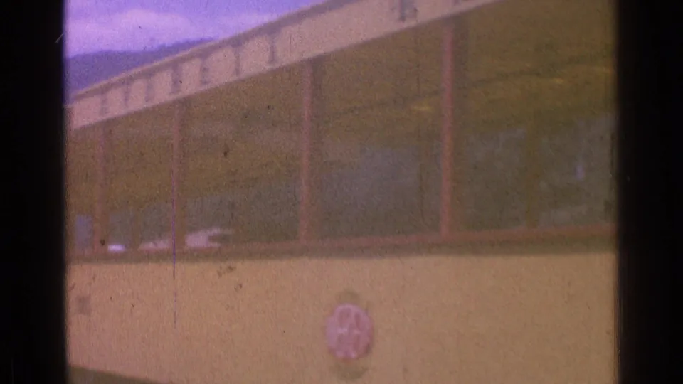 Close 1976 8mm view of a yellow train car side, showing windows, weathered color, and a small red emblem that is not readable.