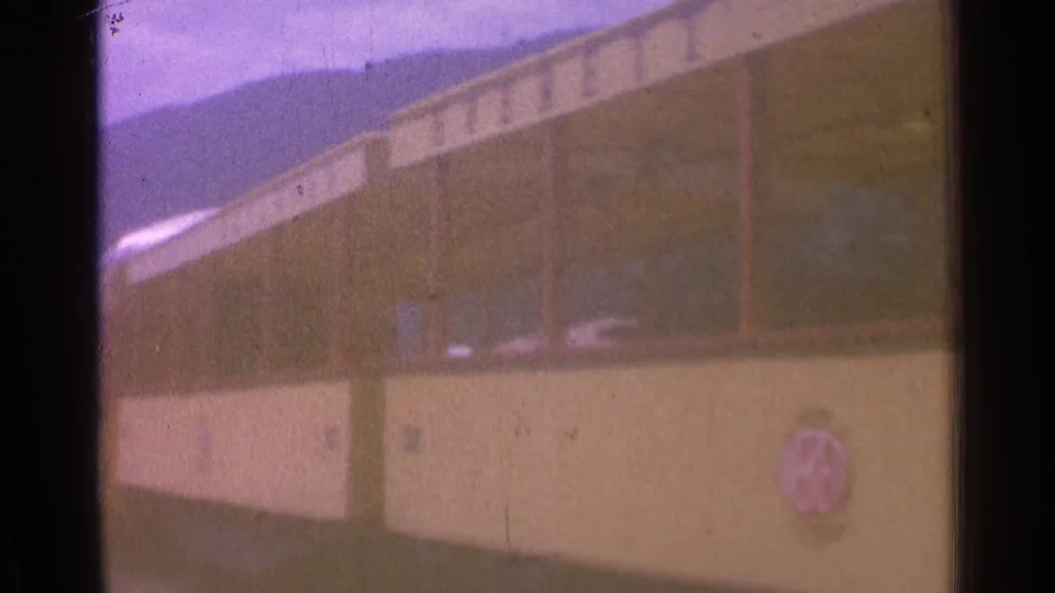 1976 8mm frame of a yellow rail car with dark windows, soft focus, and distant hills or mountains beyond the station area.