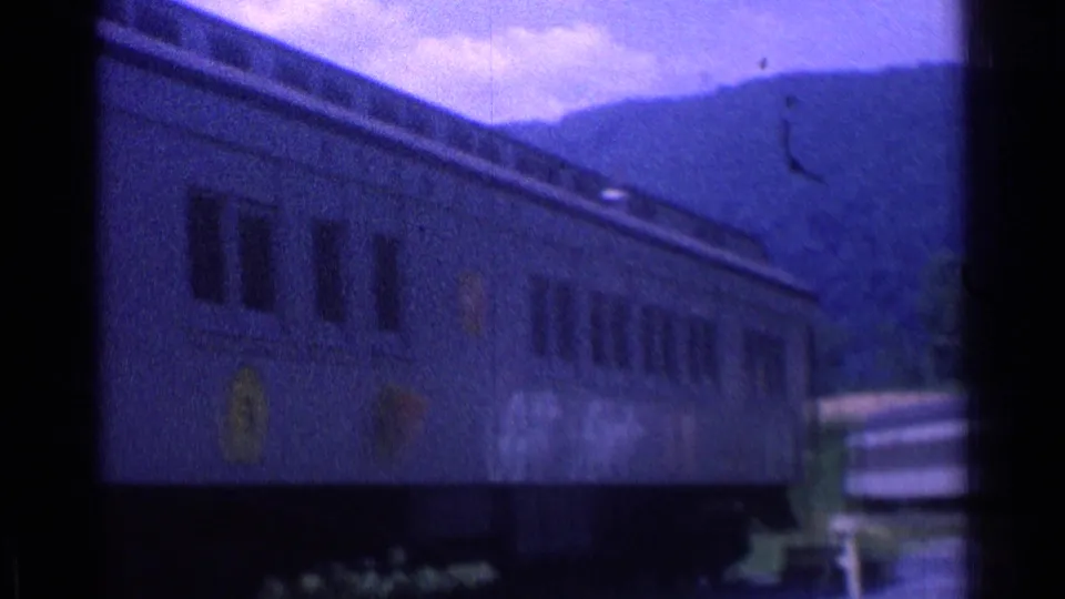 1976 home-movie frame of a stationary rail car near a small platform, with wooded hills or mountains in the background.