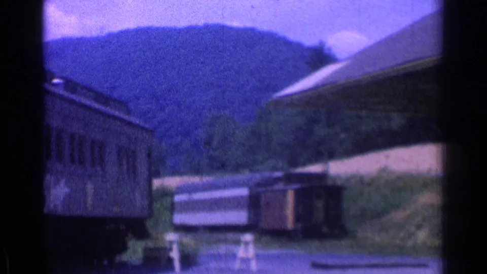 Faded 8mm frame showing the end of a rail car, a platform or station structure, and tree-covered hills beyond.