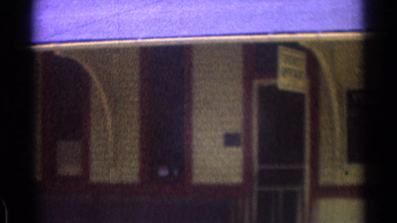 1976 8mm frame of a rail station facade with arched trim, a doorway, and a small posted sign near the entrance.