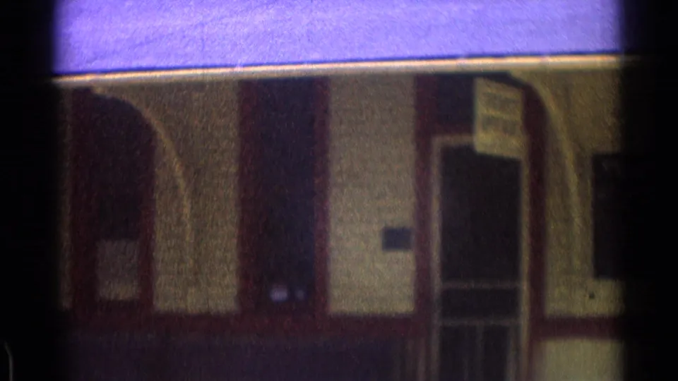 1976 8mm frame of a rail station facade with arched trim, a doorway, and a small posted sign near the entrance.