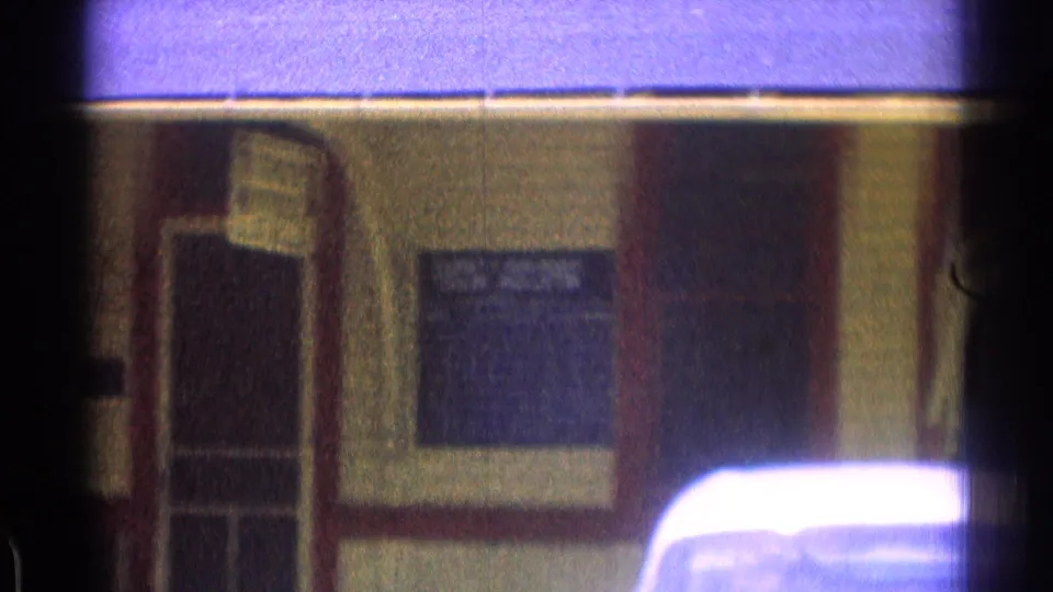 1976 8mm frame showing the station wall, a window or ticket-office sign area, and the roofline of a parked car in front.
