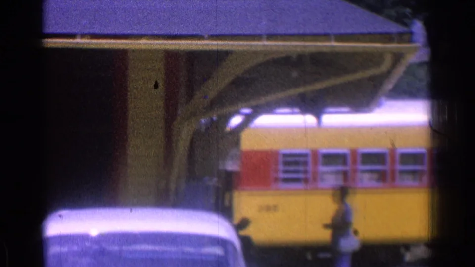 1976 8mm frame with a yellow-red passenger car beside the covered platform and one person walking near the tracks.