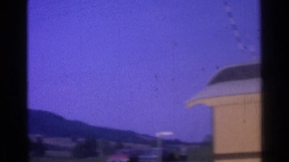1976 8mm frame of the building corner with a curved roof detail, open sky and distant landscape beyond.