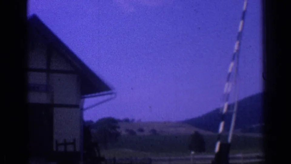 1976 8mm frame showing a dark roofed structure near distant hills, with a striped vertical pole at the right edge.