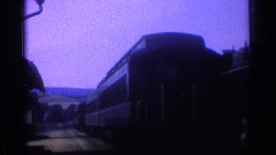 1976 8mm frame showing dark train cars along the platform, with open landscape visible beyond the station area.