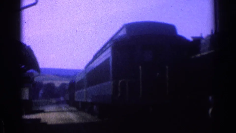 1976 8mm frame of dark train cars at a platform, with station structure fragments and countryside in the background.
