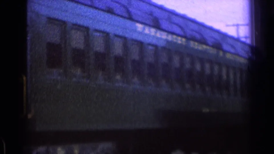 Soft 8mm view of a dark green passenger car with windows and lettering beside a station building.