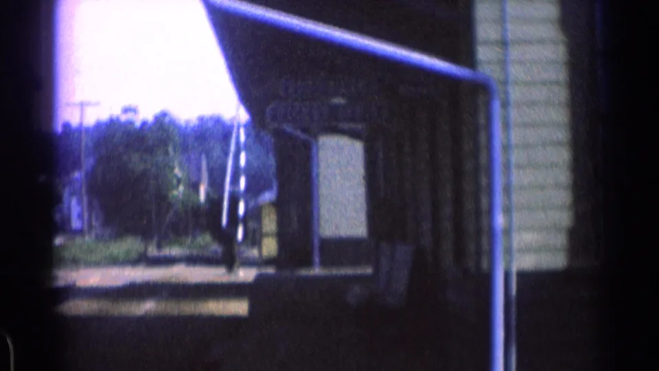 Home-movie frame looking along a small station platform, with train equipment partly visible and trees in the distance.