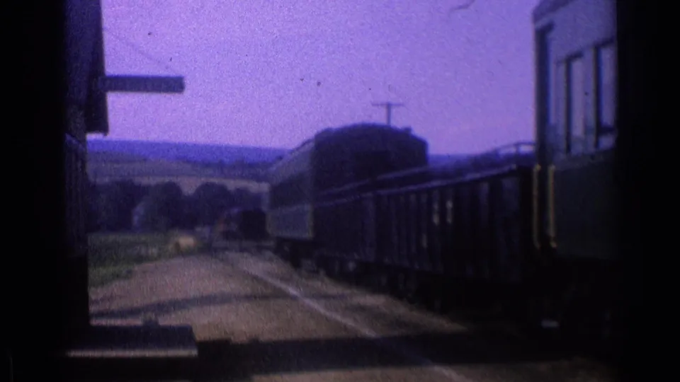Passenger rail cars and a platform sign appear in a soft 1976 8mm station view near open countryside.