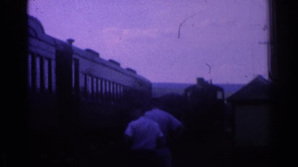 1976 8mm home-movie frame showing dark rail cars passing near a person in a light shirt, with sky and distant countryside behind.