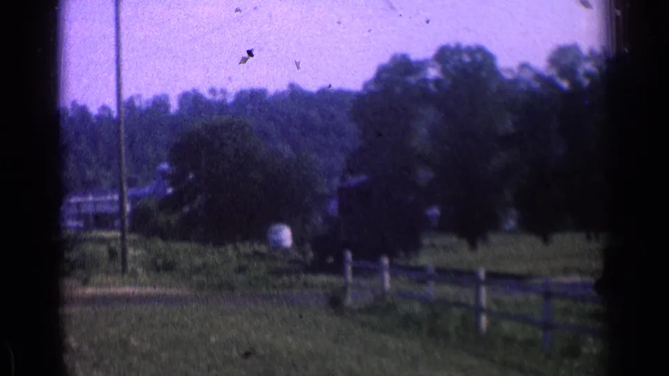 1976 8mm home-movie view of open countryside, trees, a wooden fence, utility pole, and distant farm-like structures.