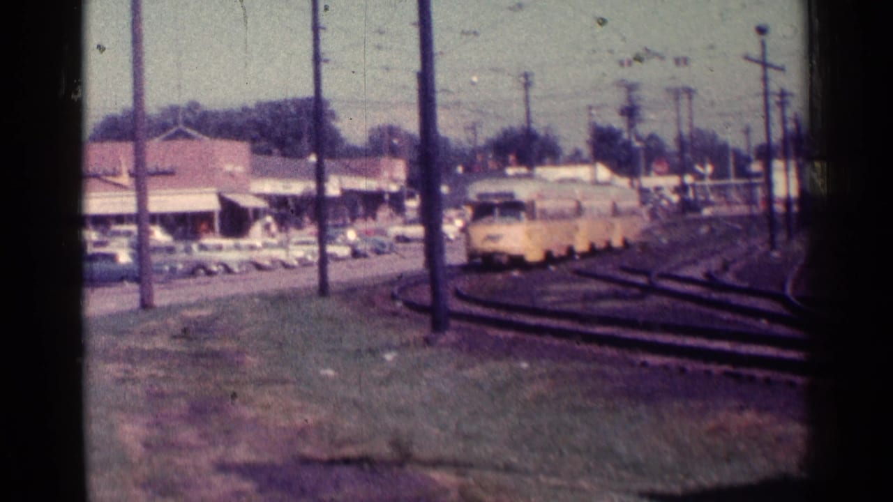 Yellow trolley approaches on curving tracks beside a parking lot and storefronts.