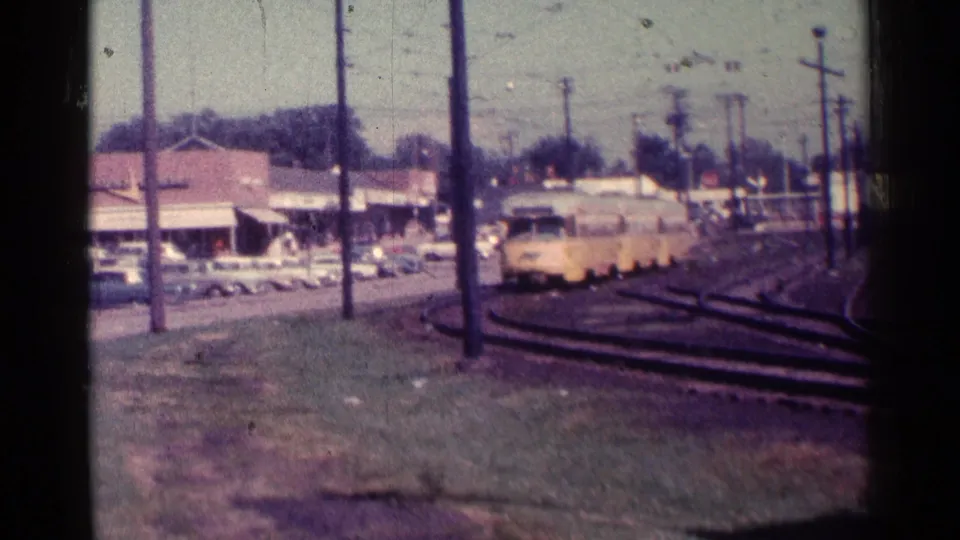 1976 Chicago 8mm frame showing a yellow trolley in the distance on rail tracks near parked cars, storefronts, utility poles, and overhead wires.