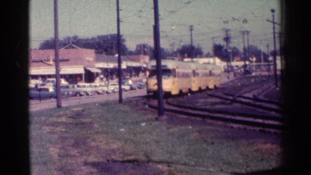 Yellow trolley moves closer along tracks near utility poles and parked cars.