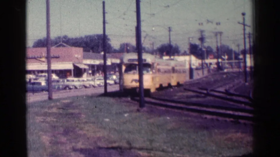 1976 Chicago 8mm frame showing the trolley close to the camera, with curved tracks, parked cars, storefront awnings, and overhead utility lines visible.