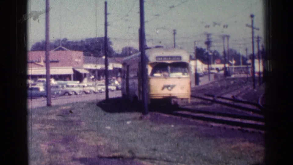 1976 Chicago 8mm frame with the front of a yellow trolley close to camera, rail tracks to the side, and storefronts, parked cars, and poles behind it.