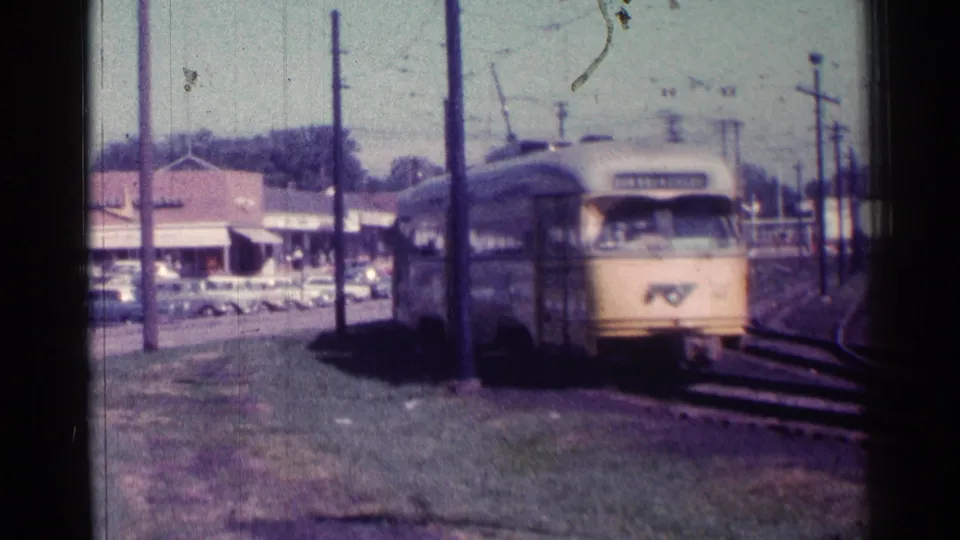 1976 Chicago home-movie frame showing a yellow rapid transit train approaching along tracks near storefronts and parked cars.