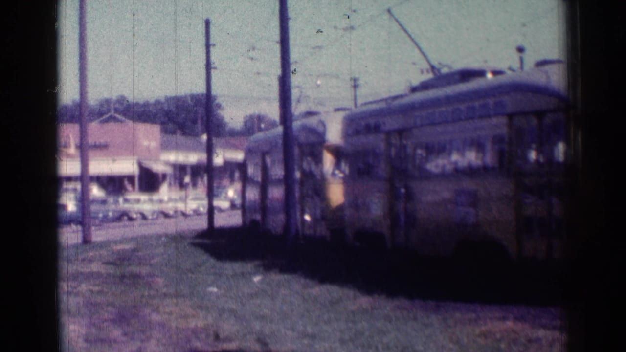 Train cars pass the camera with storefronts and parked cars in the background.