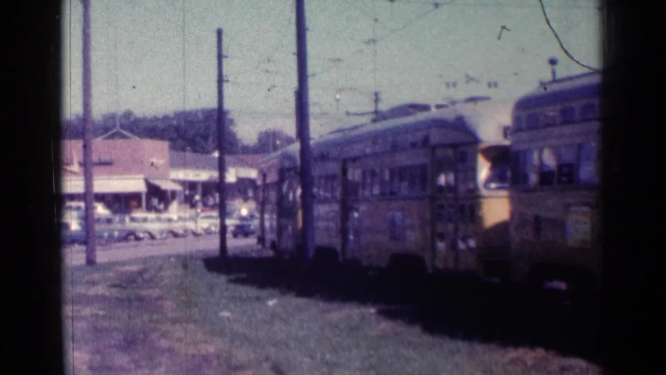 1976 Chicago home-movie view of several yellow rapid transit cars moving by, with overhead lines and storefronts beyond.