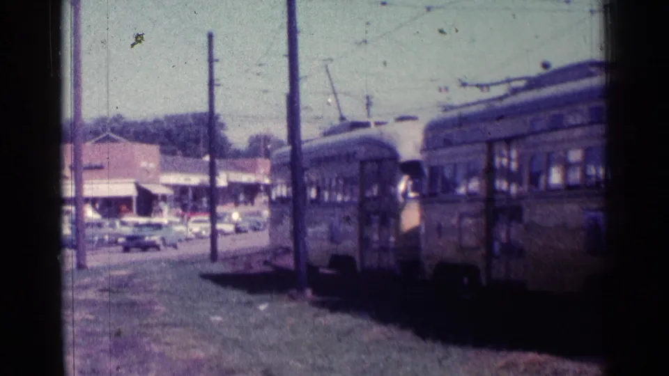 1976 Chicago frame showing the train moving away along tracks beside a street lined with cars, storefronts, and poles.