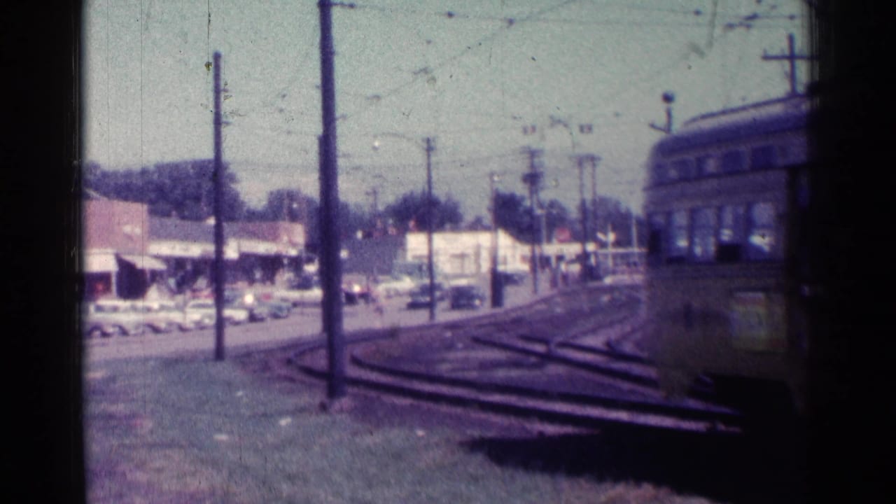 Trolley leaving frame at right with tracks, wires, storefronts, and parked cars visible.