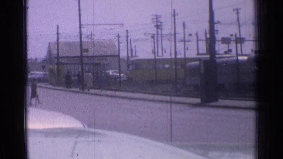 1976 Chicago home-movie frame with a distant pedestrian, rail tracks, utility poles, parked cars, and the trolley at right.
