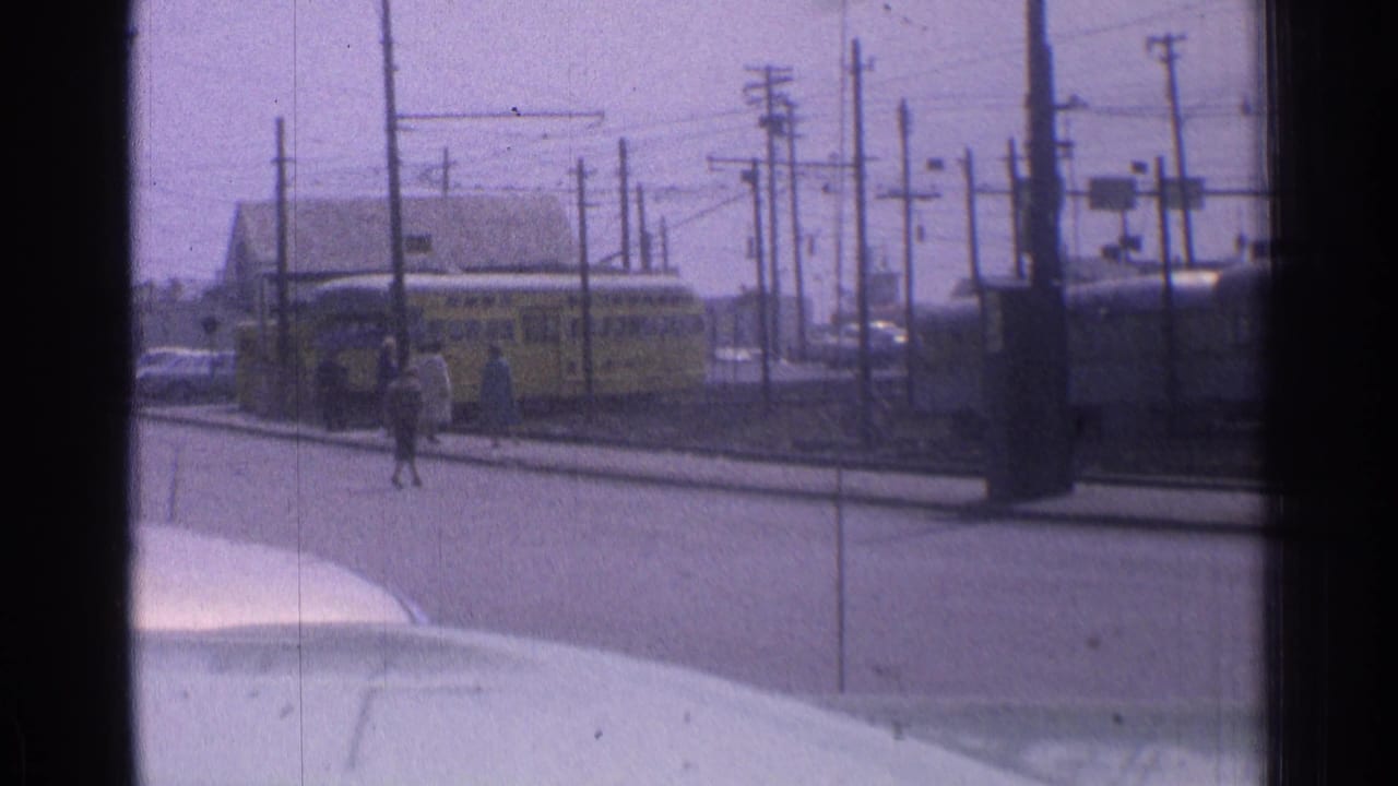 Pedestrians cross near a yellow transit vehicle on a wire-lined street.