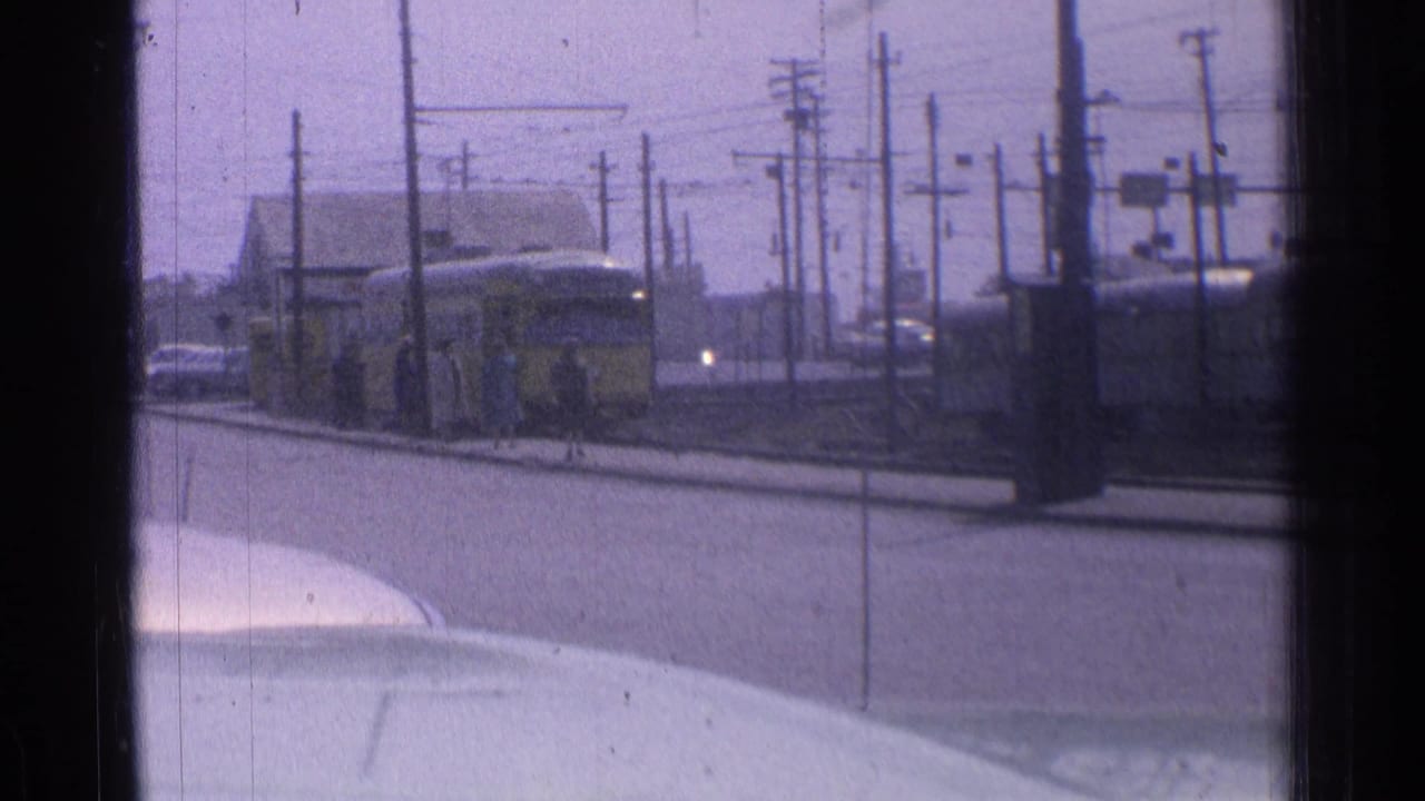 Yellow tram approaches along street tracks with utility poles and waiting passengers nearby.