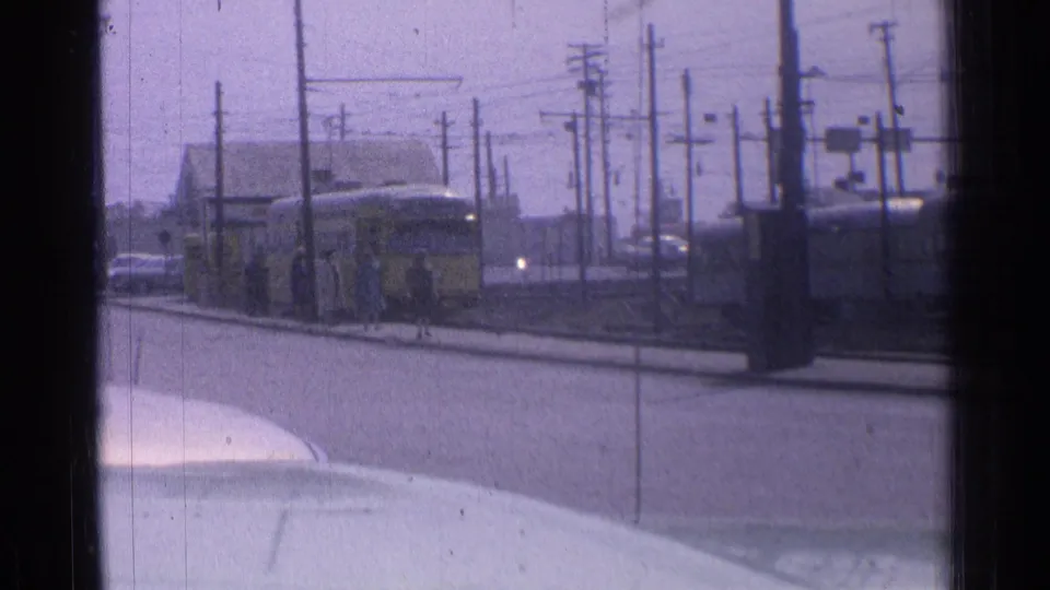 1976 Chicago 8mm frame showing a yellow tram approaching a stop beside overhead wires, poles, cars, and people waiting at the curb.