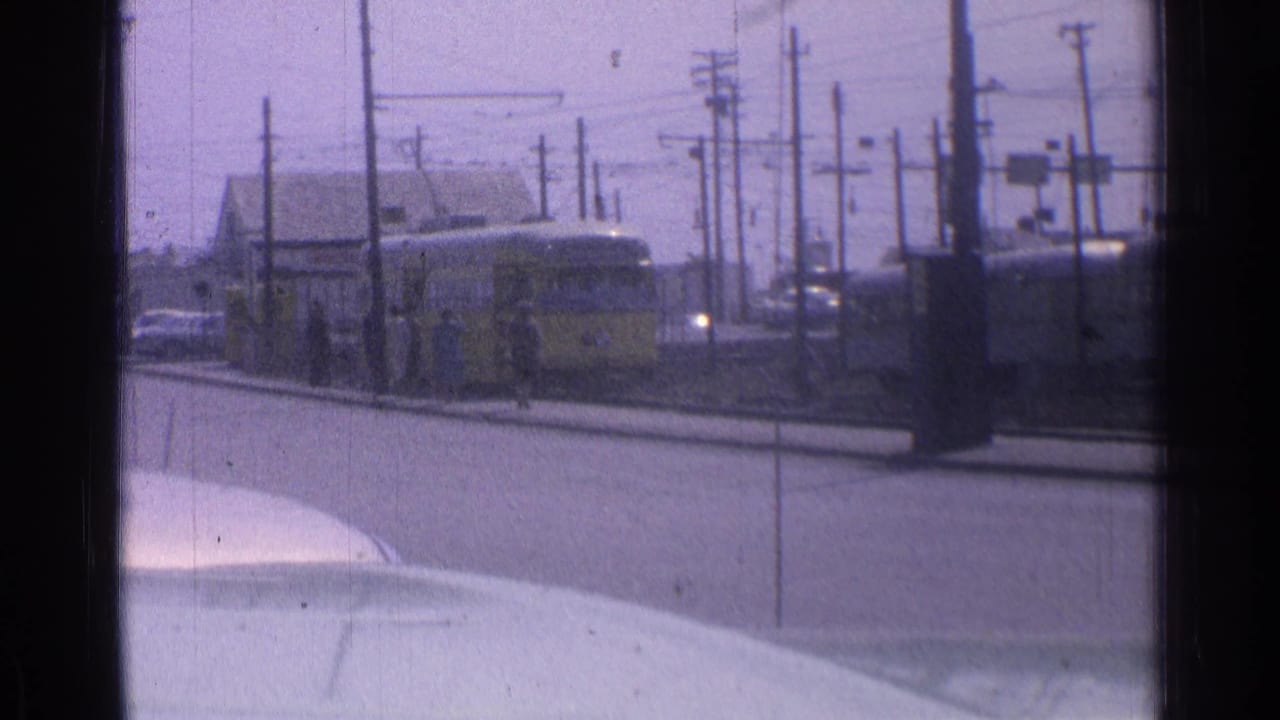 Yellow tram nears the stop on tracks under overhead wires with people beside the street.