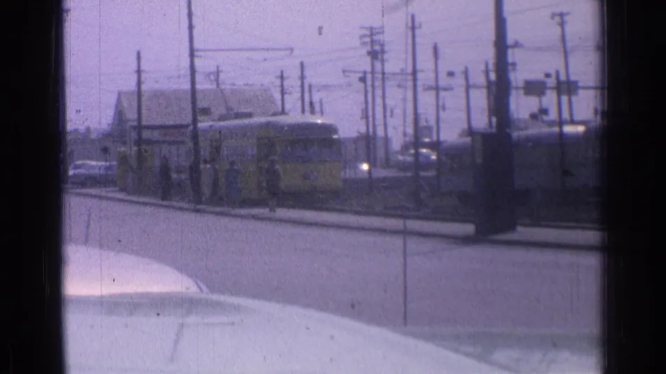 1976 Chicago transit scene with a yellow tram nearing a curbside stop, utility poles, overhead wires, cars, and distant passengers visible.